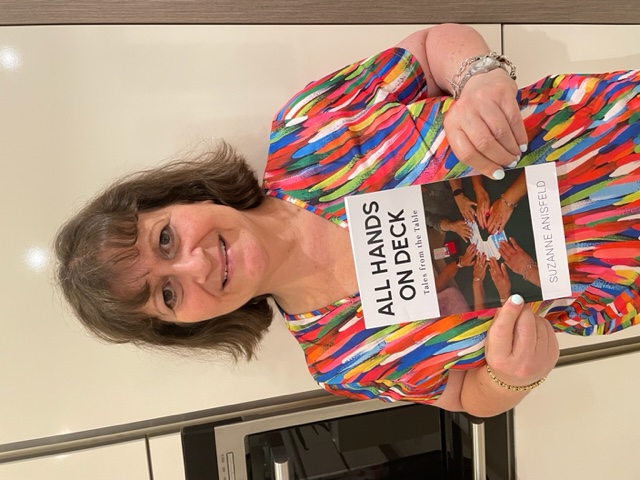 Photo of Suzanne Anisfeld, she smiling at the camera, wearing a colourful dress and holding a copy of her book titled "All hands on deck - tales from the table"