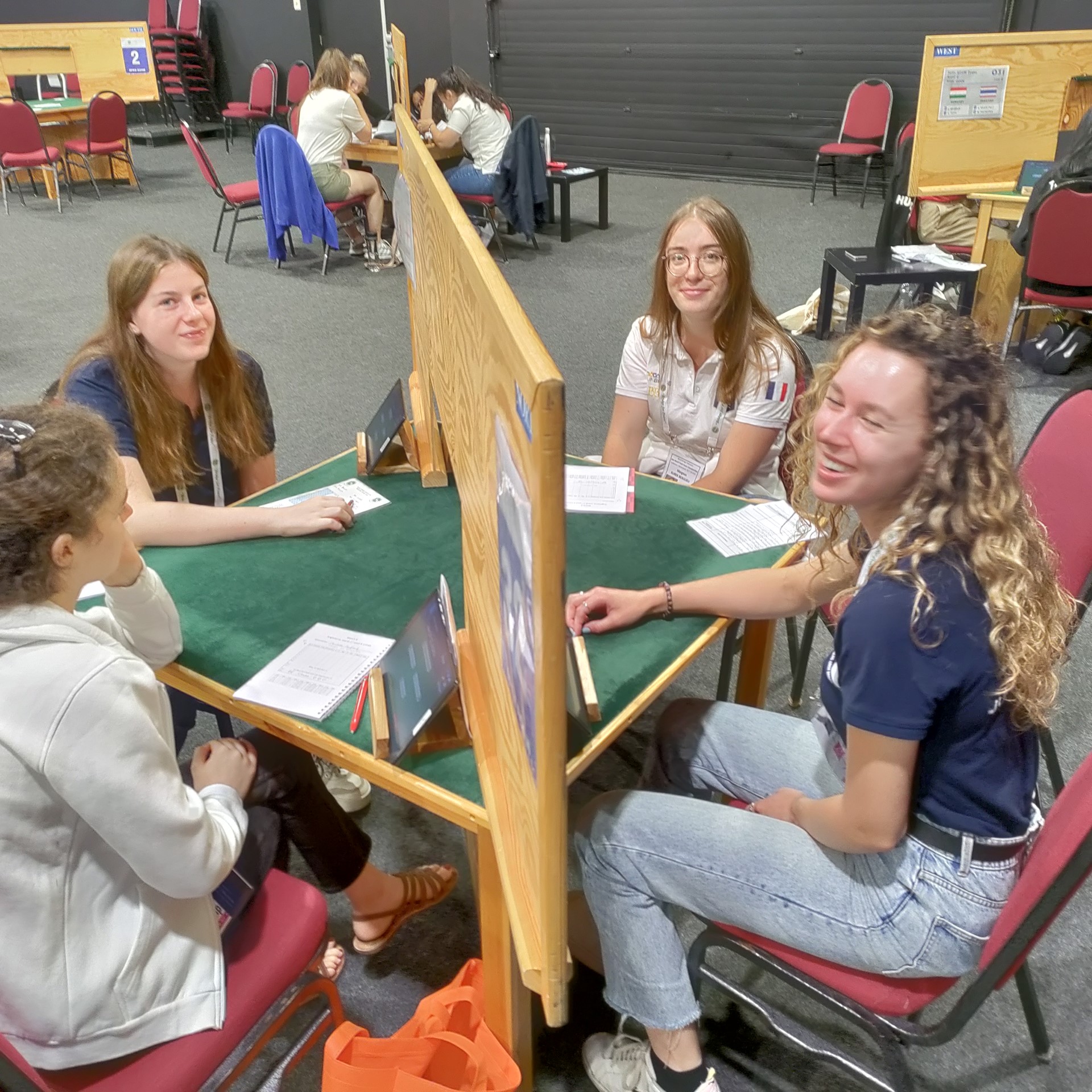 Photo of some of two of England U26 Women's team at a bridge table with two French players, the bridge table has a diagonal screen so partners cannot see each other